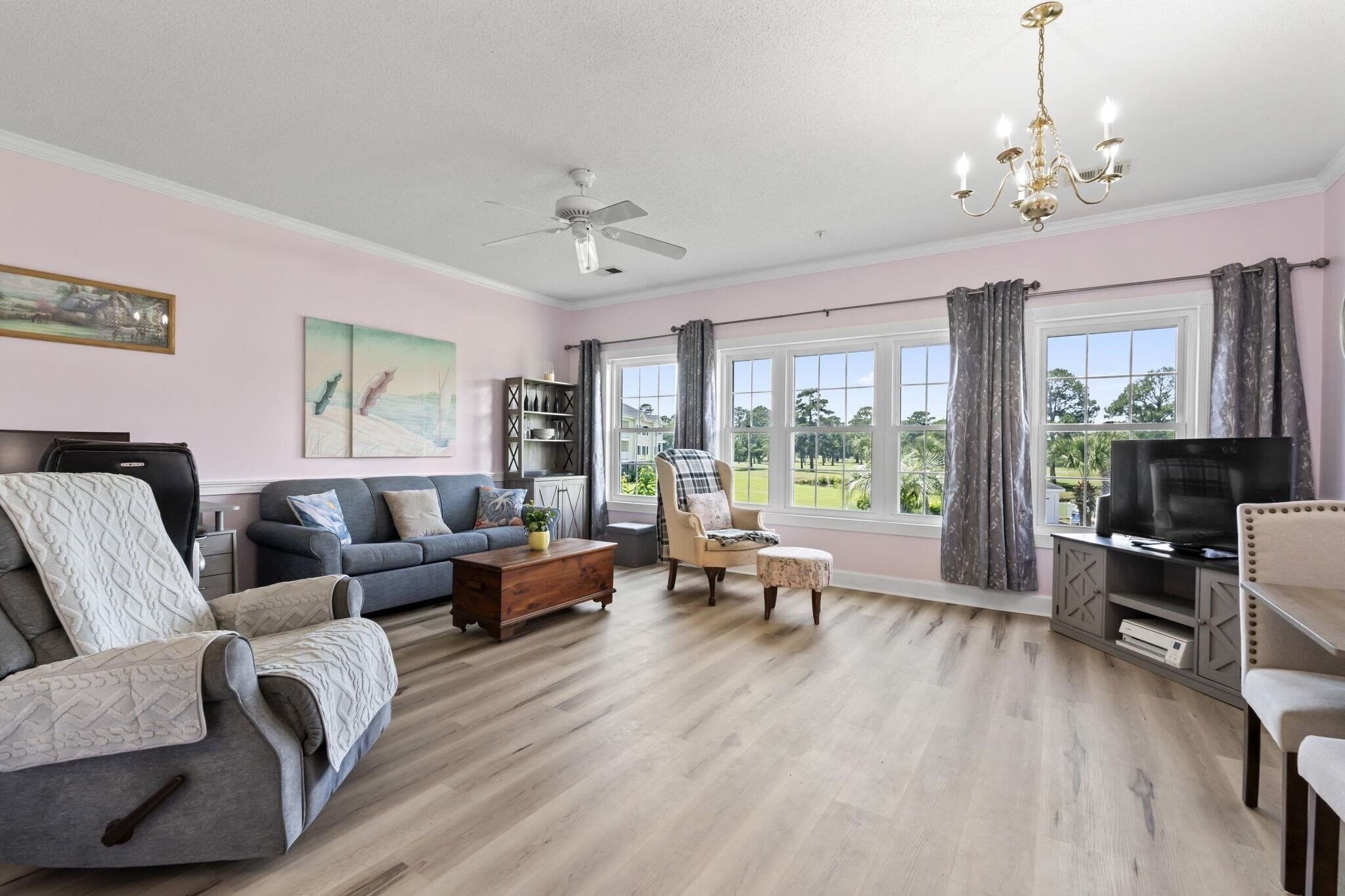 A living room with a sofa, armchair, coffee table, and a television. There are windows with curtains, a ceiling fan, and a chandelier.