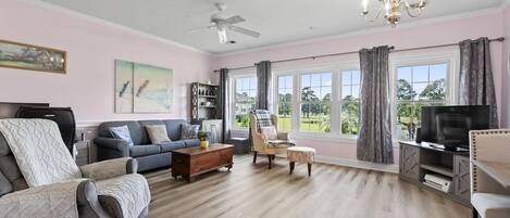 Ein Wohnzimmer mit einer Couch, einem Sessel, einem Couchtisch und einem Fernseher. Es gibt Fenster mit Vorhängen, einen Deckenventilator und eine Deckenleuchte.