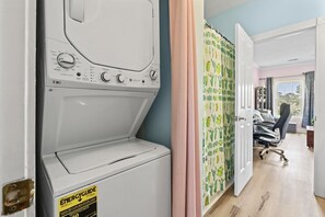 A laundry room with a front-loading washing machine, a door with a cactus-patterned curtain, and a glimpse of a bedroom with a desk and chair. - King Bed 1BR Condo | Golf View | Pool (Myrtle Beach)