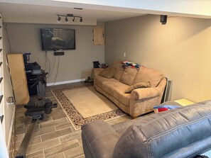 Ein Wohnzimmer mit einem beigefarbenen Sofa, einem braunen Hocker, einem Laufband und einem an der Wand befestigten Fernseher.
