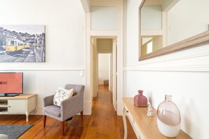 A hotel room with a wooden floor, a grey armchair, a small table with a vase, a painting on the wall, and a television on a cabinet. - GuestReady - Family home near Santa Justa Lift (Lisbon)