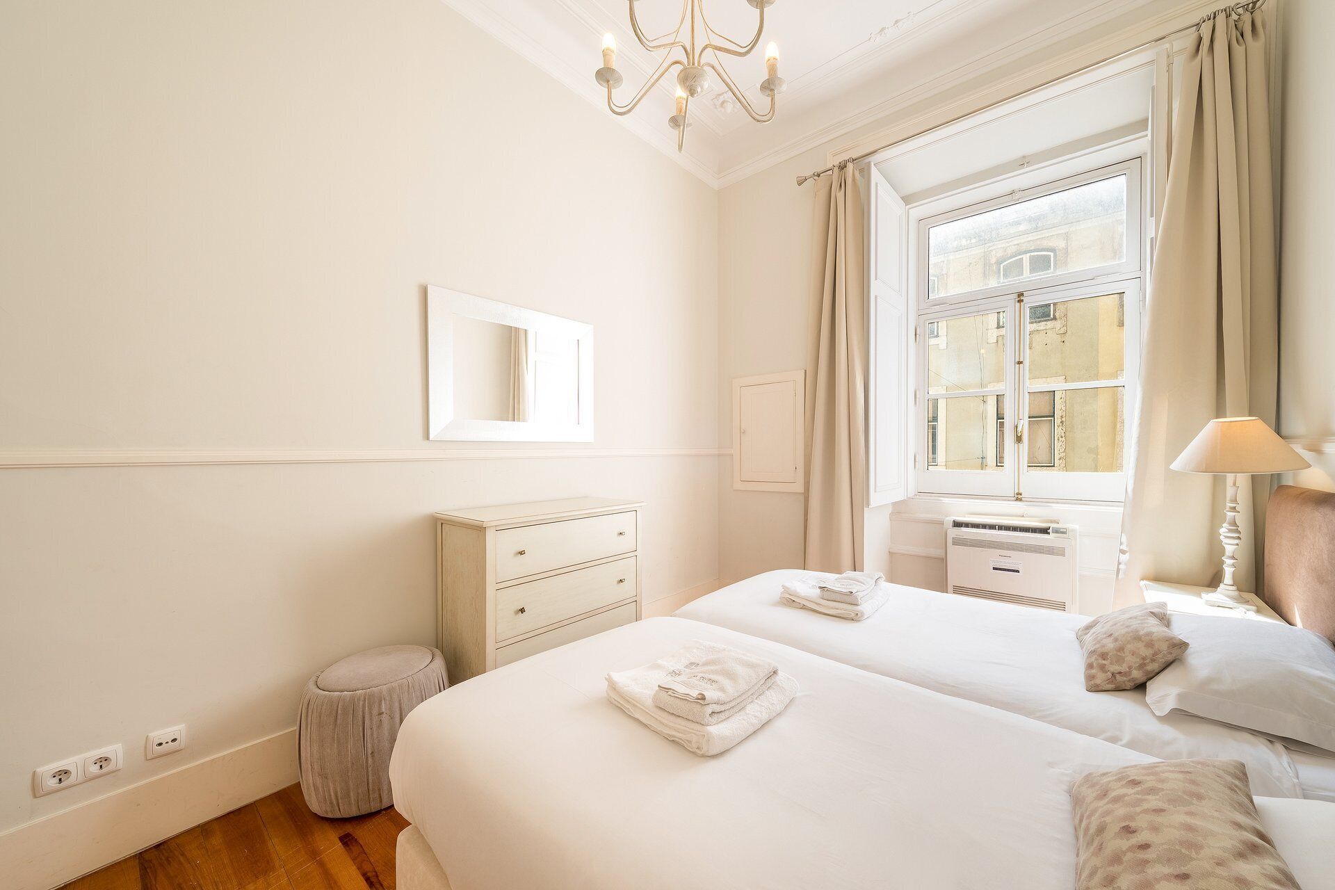 Een hotelkamer met twee bedden, een raam met gordijnen, een radiator, een bedlampje en een spiegel aan de muur.