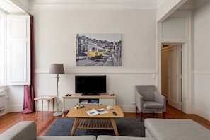 A modern living room with a flat-screen TV, a coffee table, a sofa, a chair, and a painting of a tram on the wall. - GuestReady - Family home near Santa Justa Lift (Lisbon)