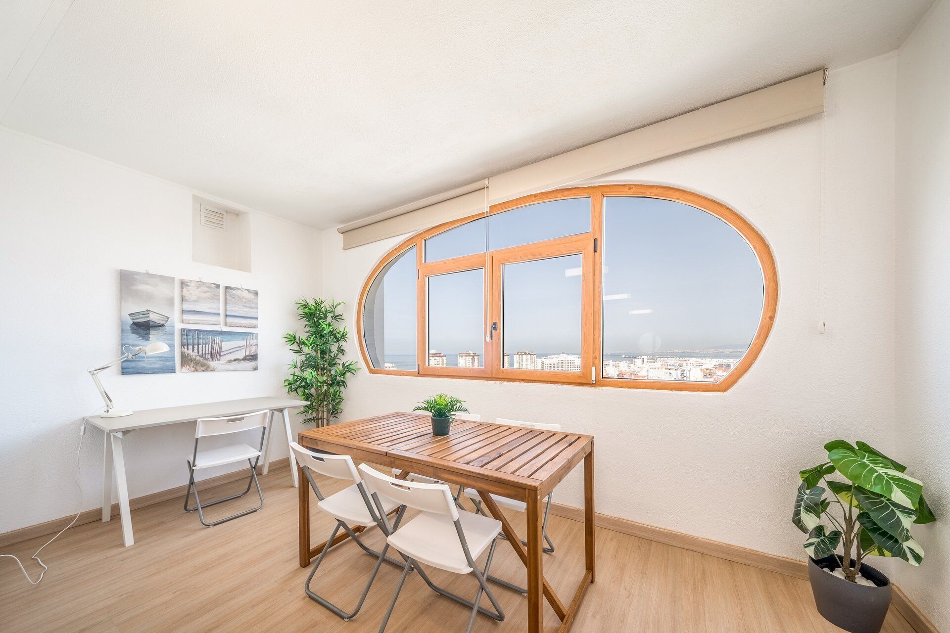 A room with a wooden table and chairs, a potted plant, and a large window with a view of buildings.