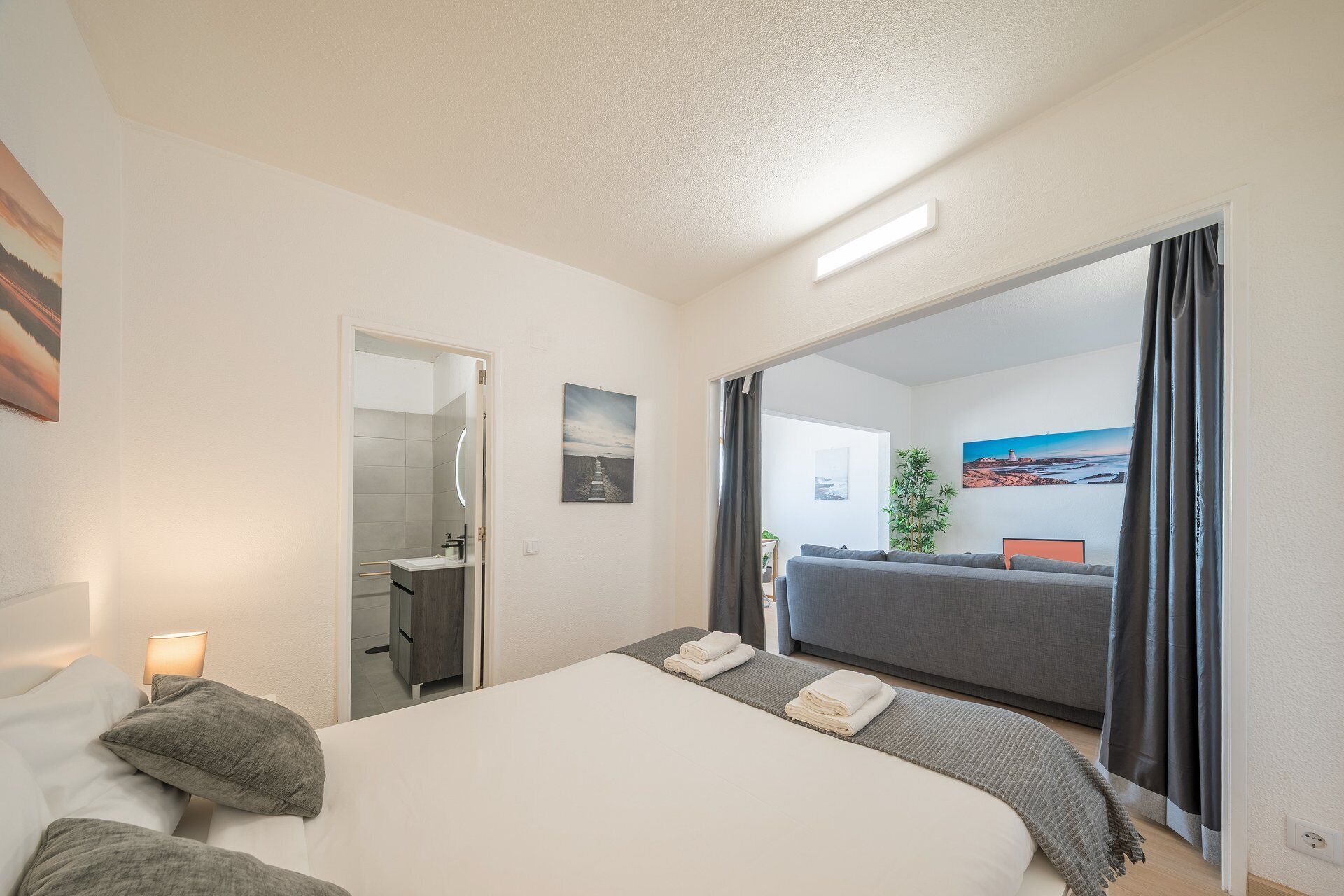 Ein Hotelzimmer mit Bett, Nachttischlampe, Badezimmer mit Spiegel und Meerblick durch ein großes Fenster.