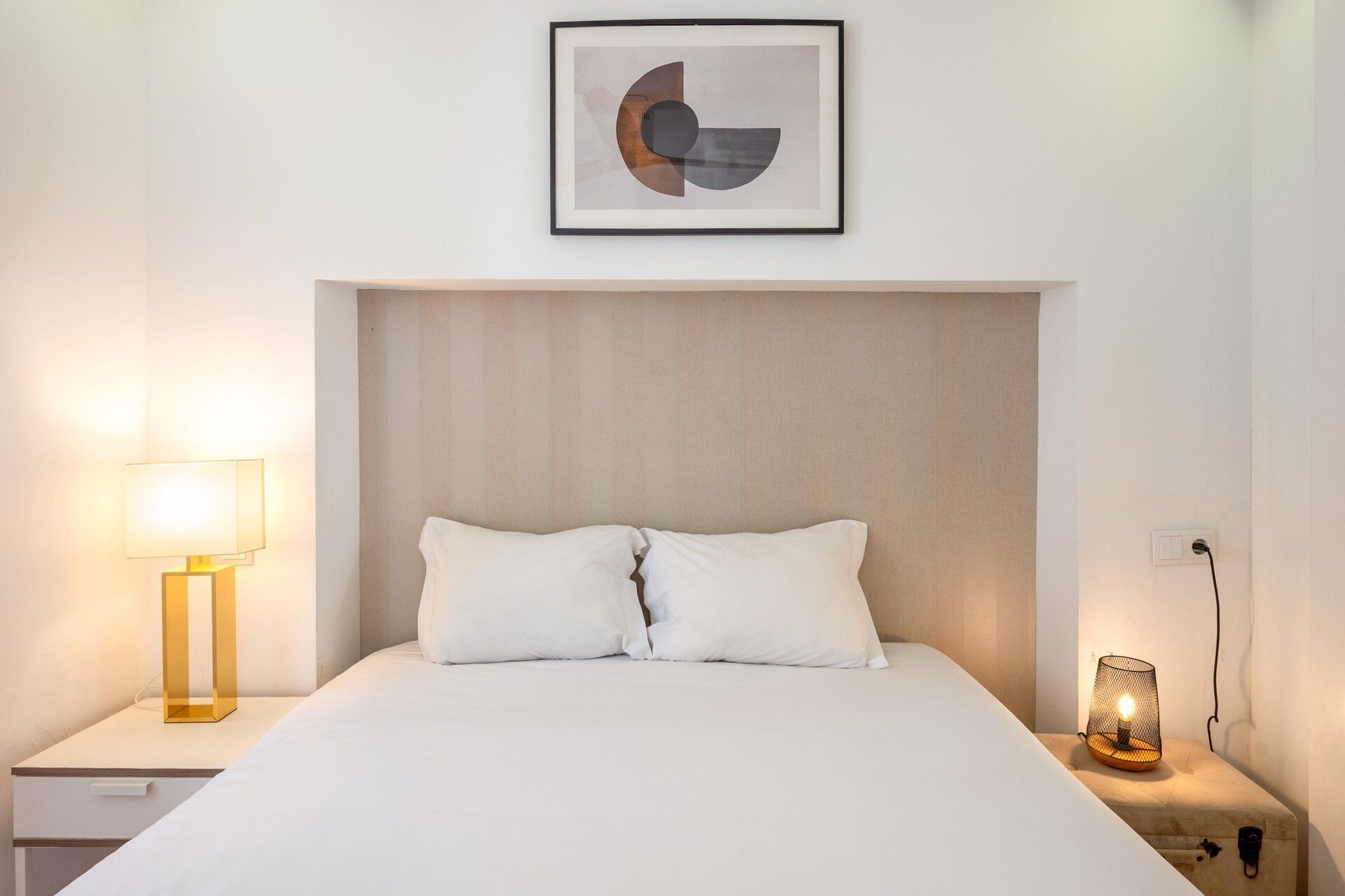 Een netjes opgemaakt bed met twee kussens, een bedlampje, een abstract schilderij aan de muur en een nachtkastje met een lamp.