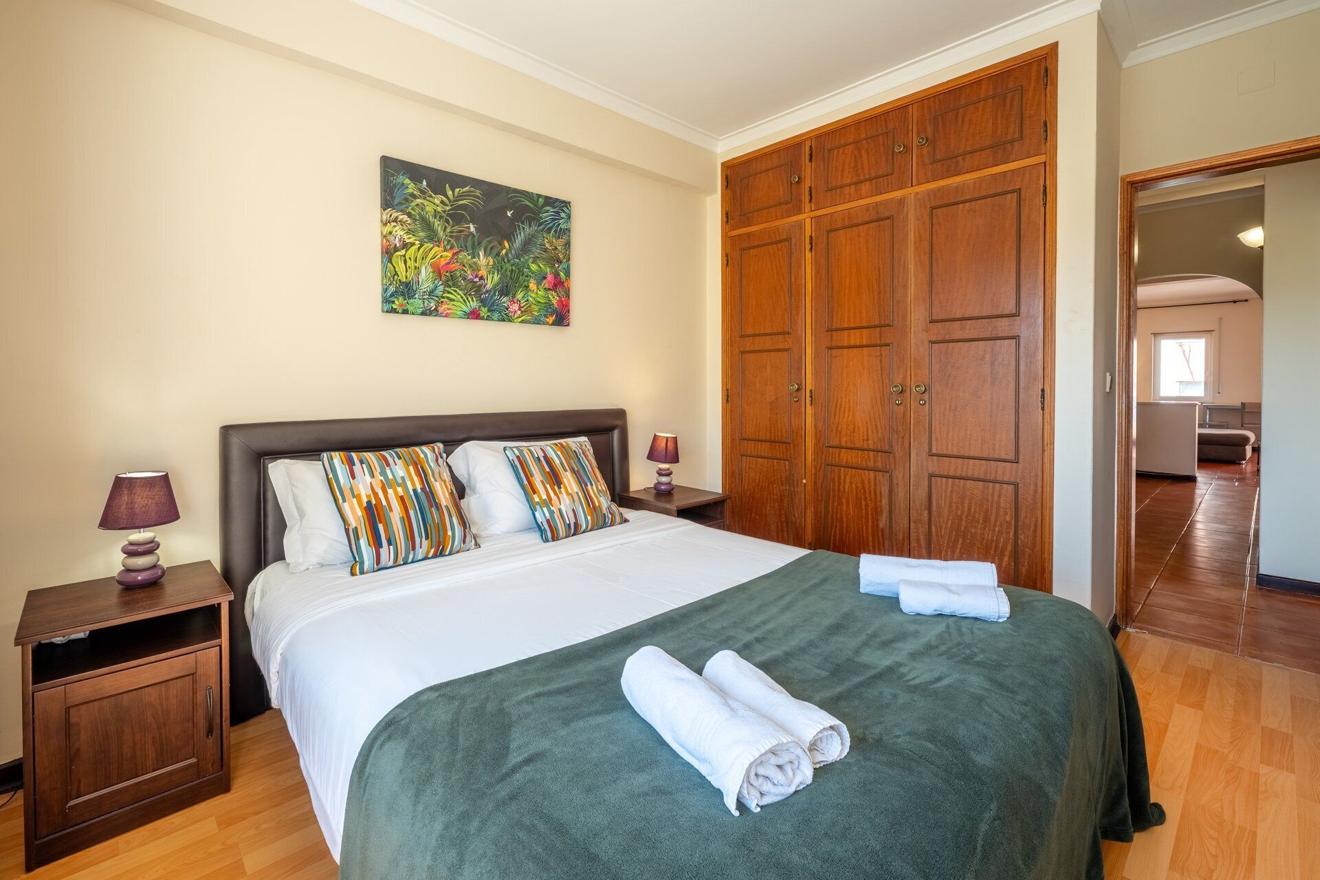 A hotel room with a large bed, wooden wardrobe, and a painting on the wall.