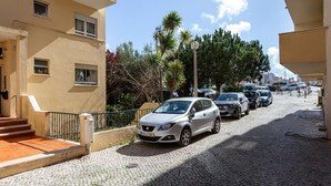 Una calle con coches aparcados, un edificio con balcón y cielo despejado.