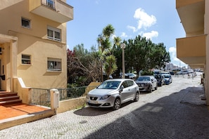 Una calle con coches aparcados, un edificio con balcón y cielo despejado.