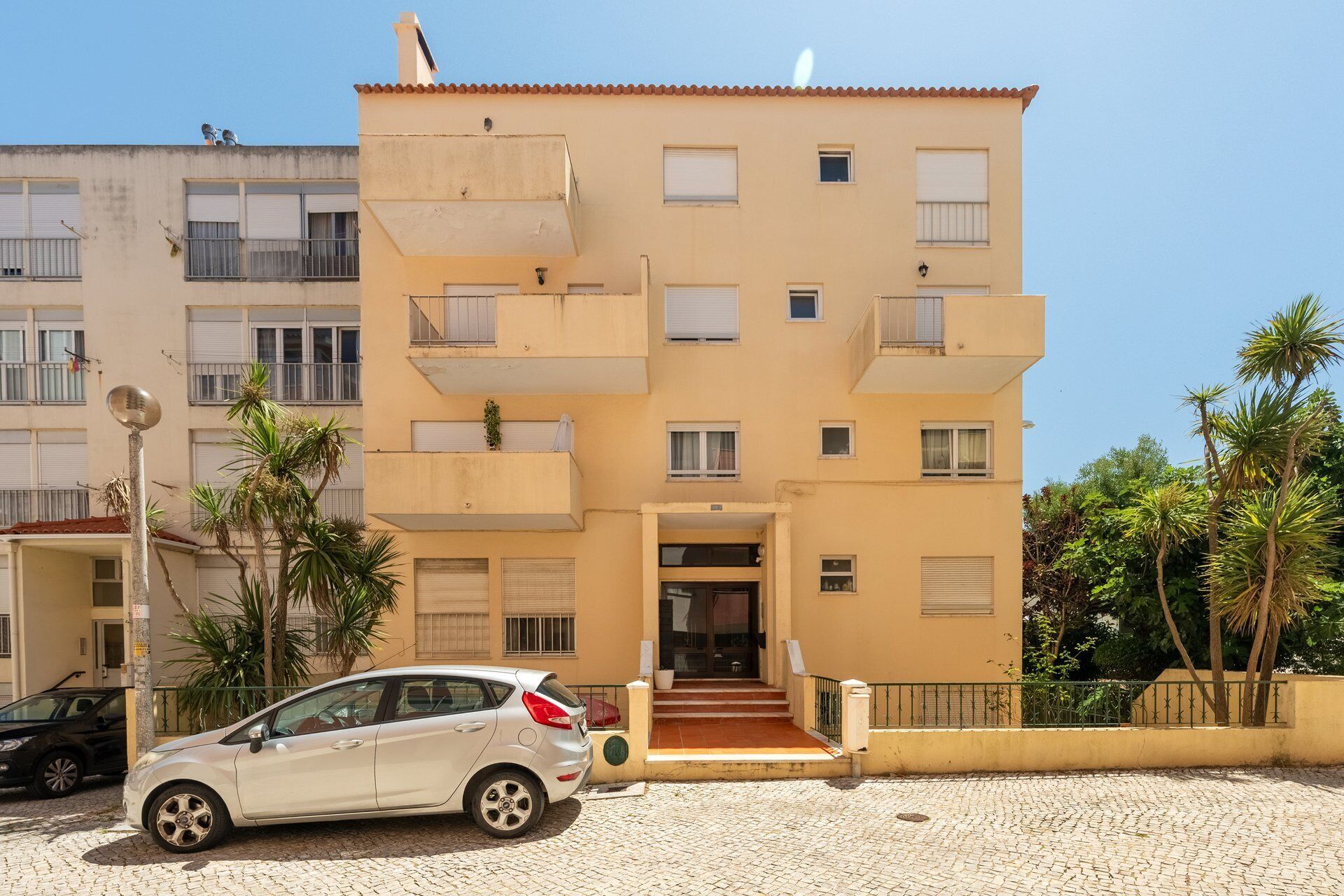 A multi-story residential building with a central entrance, balconies, and a paved ground area.