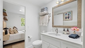 A modern bathroom with a white sink, a large mirror, and a toilet. There are towels stacked on a rack and a small shelf with toiletries. - The Big Yellow House Loft C (Summerland)