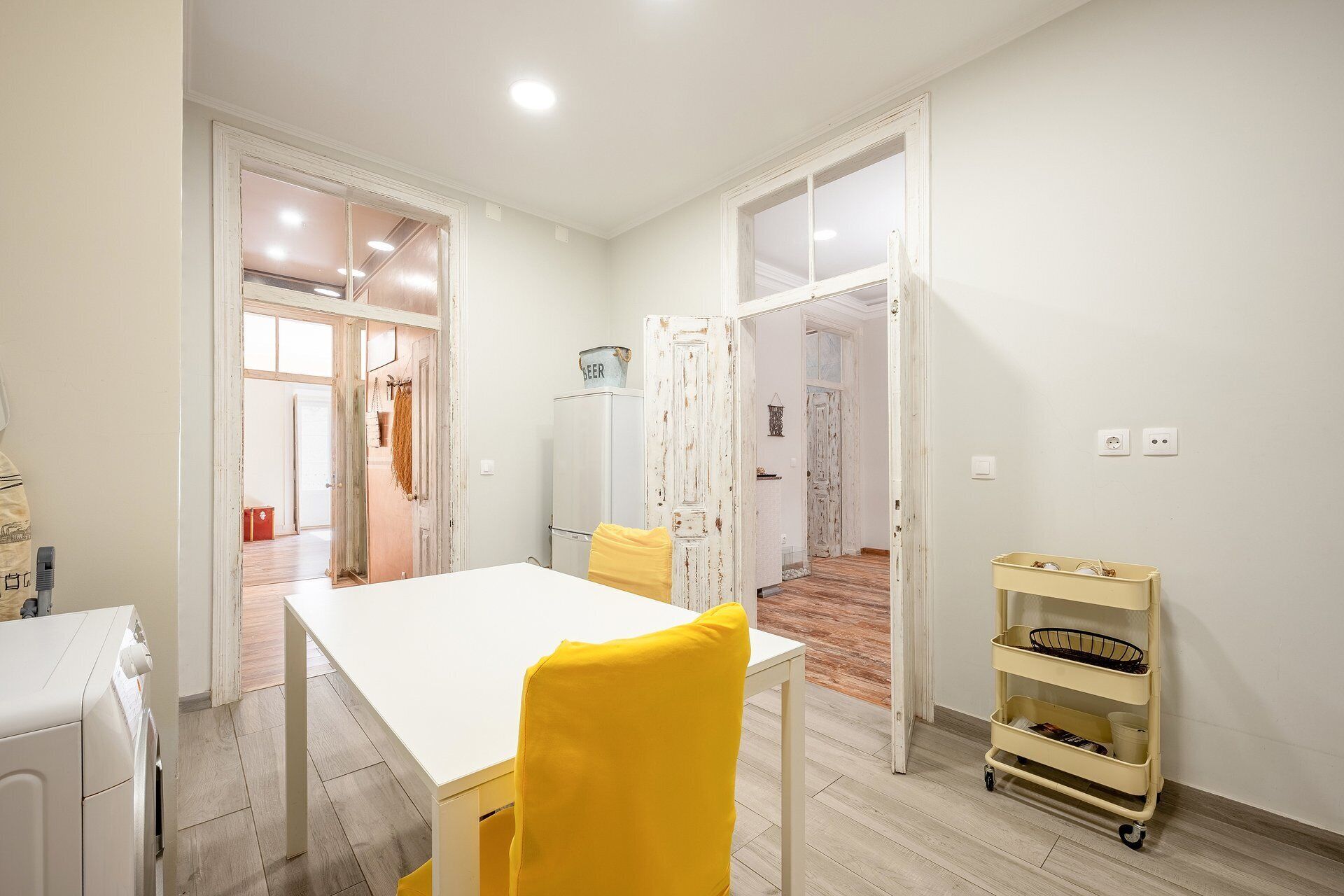 Una habitación con una mesa blanca, una silla amarilla y un piso de madera. Hay una pared blanca con una puerta que conduce a otra habitación.