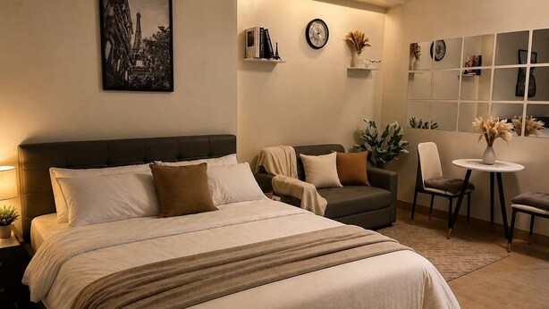 Quarto de hotel com cama, sofá, cadeira, mesa pequena e prateleira na parede com livros e relógio.