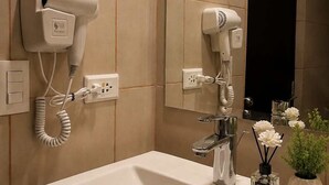 Casa de banho com lavatório branco, dois secadores de cabelo fixados na parede, espelho e planta em vaso com flores brancas.