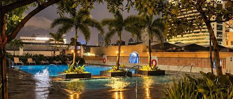 A swimming pool area with a wooden deck, surrounded by tropical plants and palm trees.