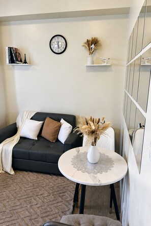 Een woonkamer met een zwarte bank, een ronde tafel met een witte vaas, een klok aan de muur, en een spiegel met een rasterpatroon.