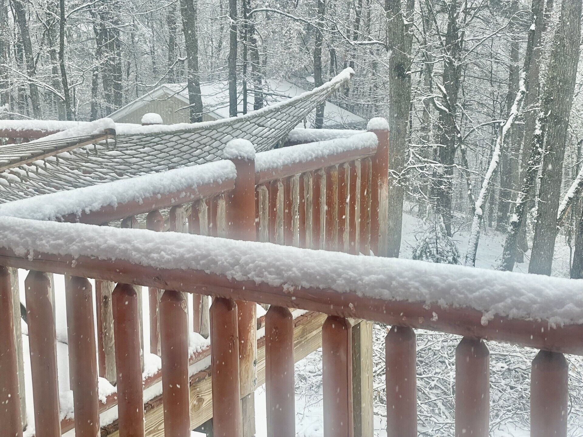 A wooden balcony railing with snow accumulation, overlooking a traditional building with a tiled roof, set in a snowy forest.