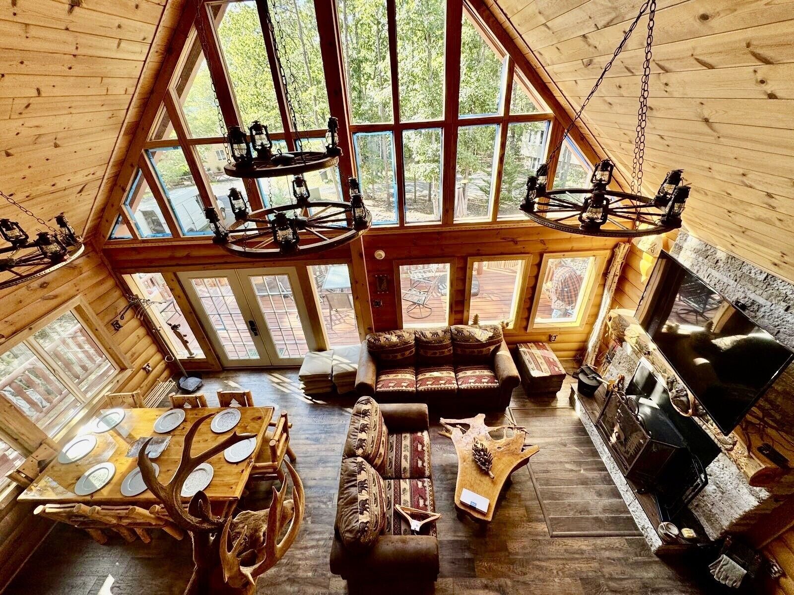 A log cabin interior with a high ceiling, large windows, a fireplace, a dining table with antler chairs, and a sofa.