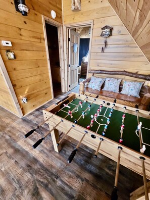 A wooden-floored room with a foosball table, a wooden bench with pillows, and a door leading to another room.