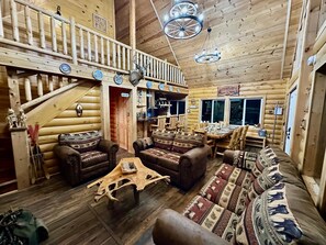 A log cabin interior with a dining area, a kitchen, and a staircase leading to an upper level.
