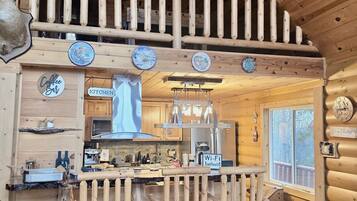 Interior rústico de uma cabana, com cozinha, área de jantar e uma varanda com guarda-corpo de madeira.