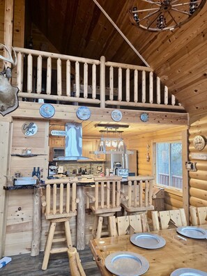 Interior rústico de una cabaña con cocina, comedor y balcón con barandilla de madera.