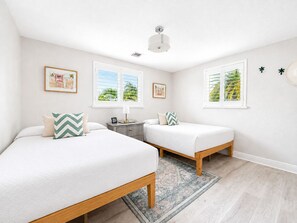 A bedroom with two beds, a nightstand, a lamp, and a view of palm trees through the windows. - Pool & Hot Tub near Siesta Key Beach (Sarasota)