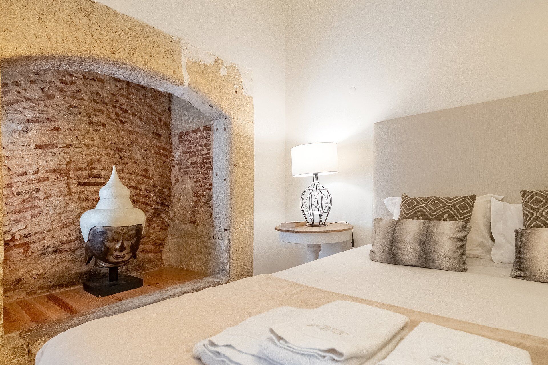 Une chambre avec une cheminée en briques, une lampe de chevet, un lit avec plusieurs oreillers et une petite table ronde.