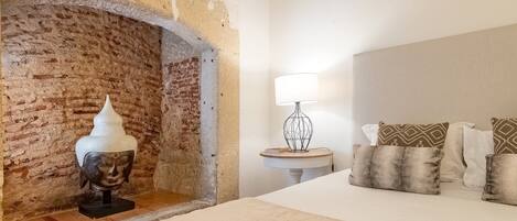 Une chambre avec une cheminée en briques, une lampe de chevet, un lit avec plusieurs oreillers et une petite table ronde.