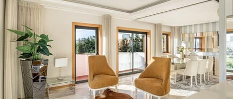 Un salón moderno con una mesa de centro de vidrio, dos sillones, una planta grande en maceta y vistas al exterior.
