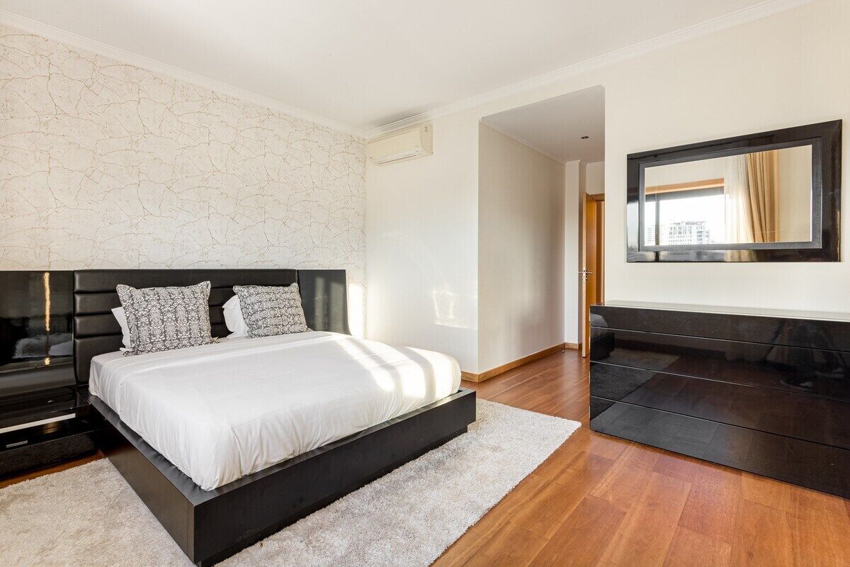 Un dormitorio moderno con cabecera negra, una cama grande, alfombra blanca y una consola negra.