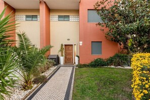 A building with a red facade, a paved entrance, and a small garden with yellow flowers. - GuestReady - Carnaxide elegant villa with 2 pools (Carnaxide)