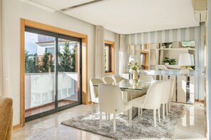 A modern dining area with a glass table and white chairs, a large sliding glass door leading to a balcony, and a striped wall with a mirror and shelves. - GuestReady - Carnaxide elegant villa with 2 pools (Carnaxide)