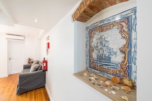 Ein Zimmer mit einer Fliesenwand, die ein Schiff und Meeresschalen zeigt, einem grauen Sofa, einem Feuerlöscher und einer weißen Tür.
