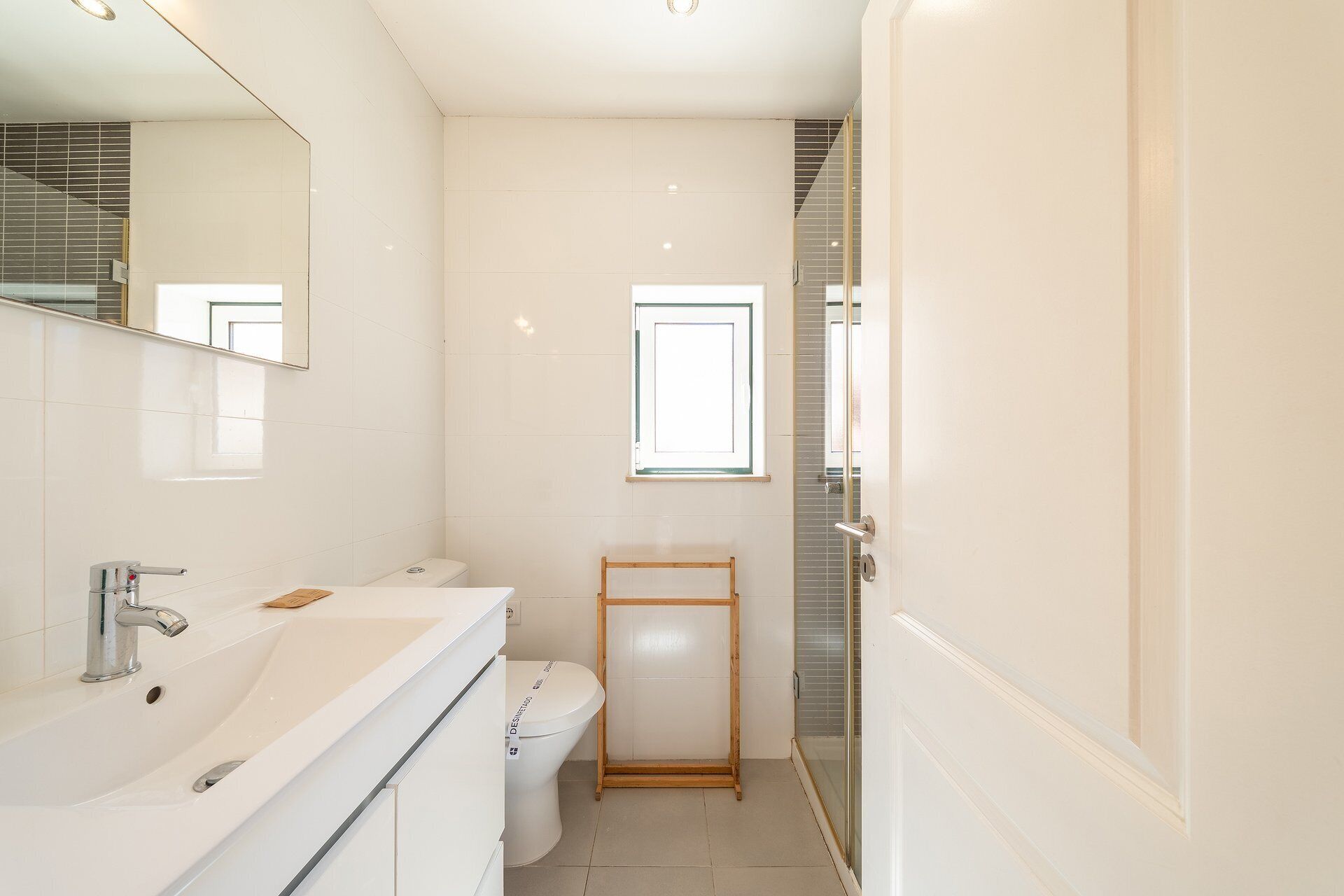 Ein modernes Badezimmer mit einem weißen Waschbecken, einem Spiegel und einer Toilette. Über der Toilette befindet sich ein Fenster und ein Duschbereich mit einer Glastür.
