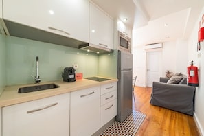 A compact kitchen with a sink, a coffee maker, a microwave, and a refrigerator. There is a grey sofa and a fire extinguisher in the adjacent living area. - GuestReady - Brilliant stay near São Jorge Castle (Lisbon)