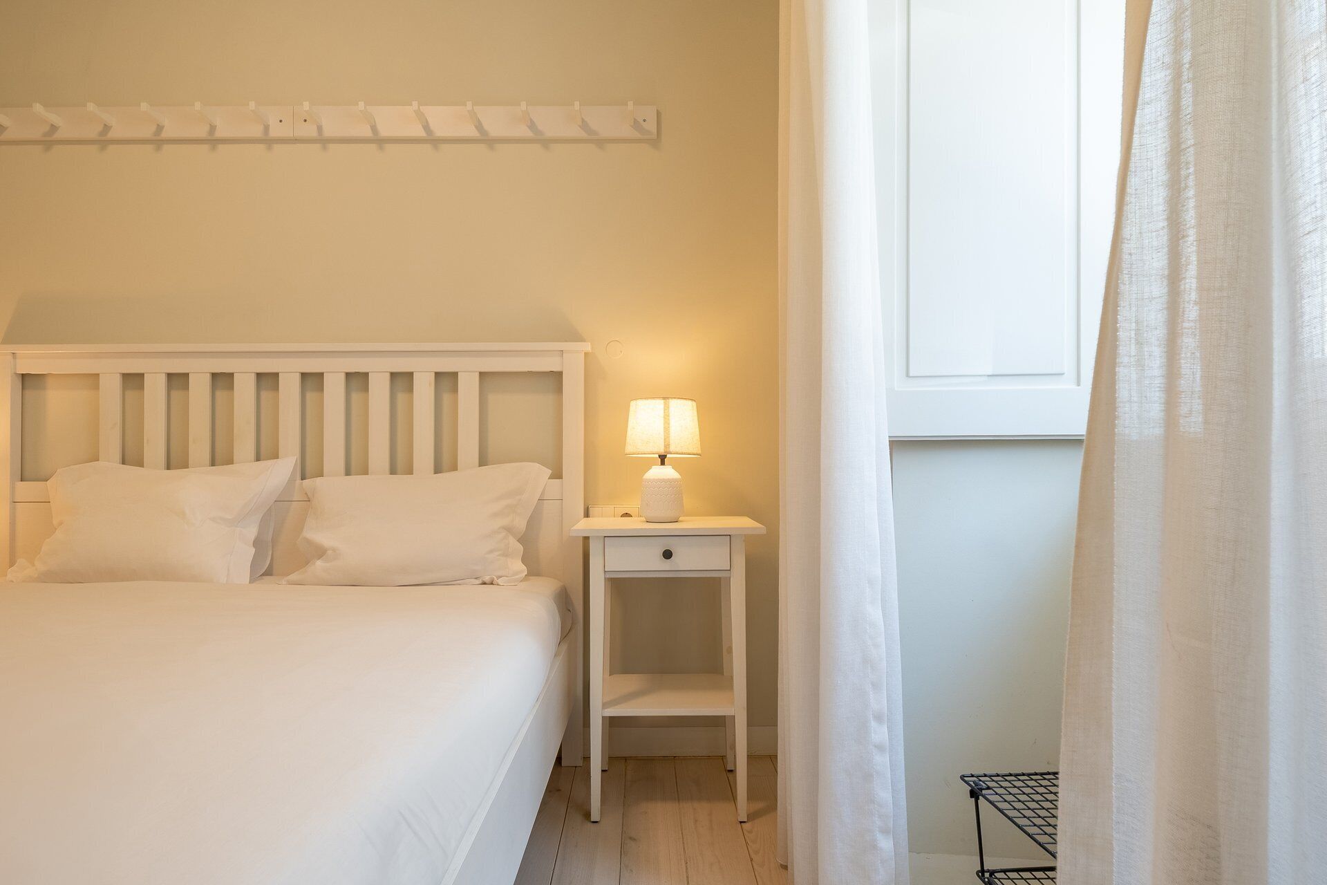 Cama com roupa de cama branca, mesa de cabeceira com abajur e janela com cortinas.