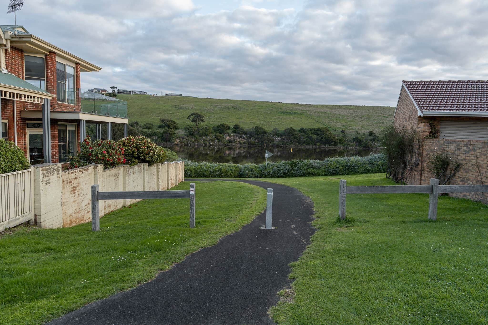 A pathway leading to a residential area with a house on the left and a hill in the background.