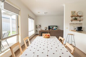 A dining area with a table set for six, a television in the background, and a kitchen area visible to the right.