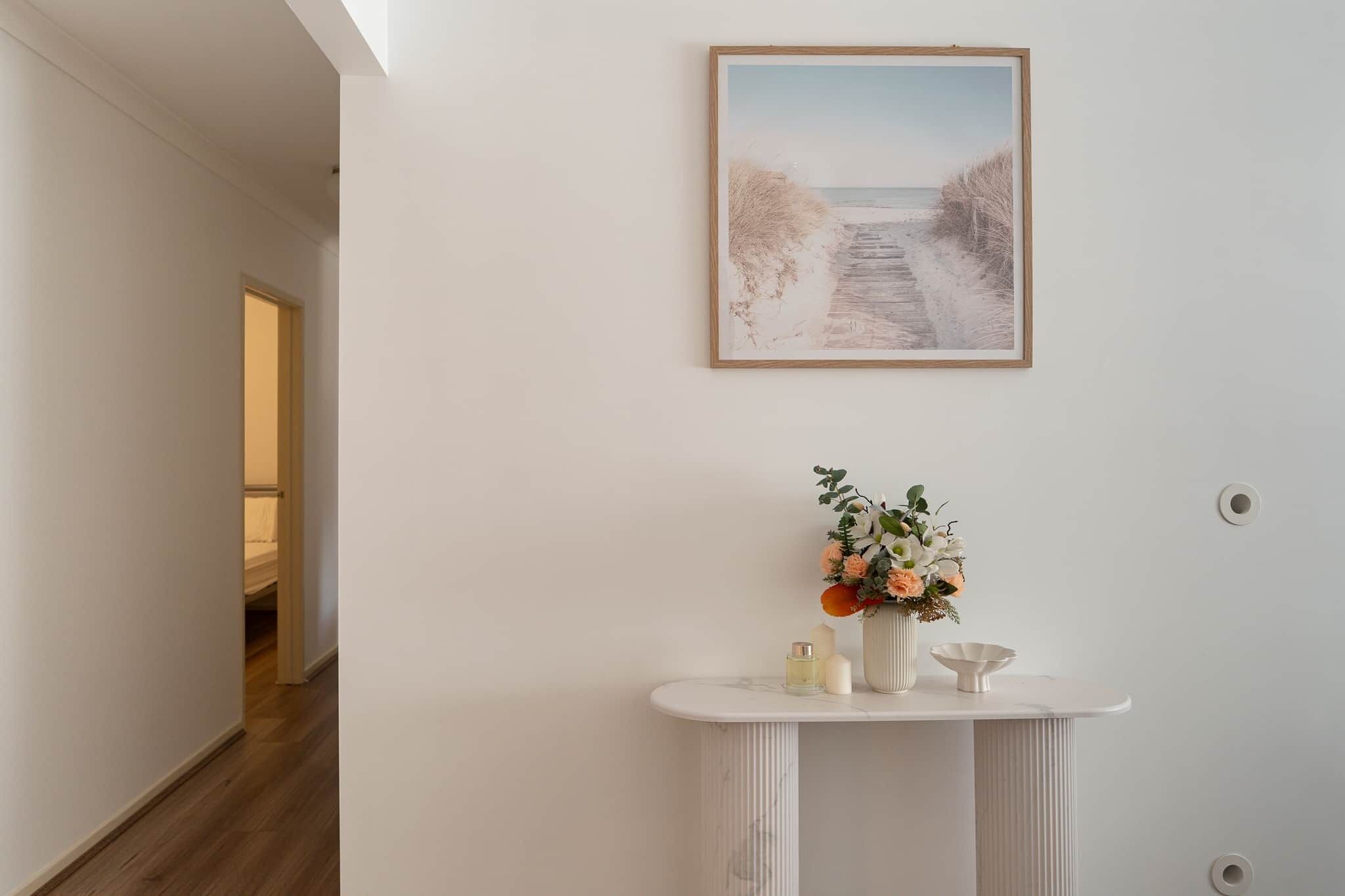 Ein Flur mit weißer Wand, einem gerahmten Bild von einem Strand und einem weißen Konsolentisch mit Blumen in einer Vase.
