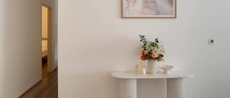 A hallway with a white wall, a framed picture of a beach, and a white console table with a vase of flowers.