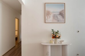 A hallway with a white wall, a framed picture of a beach, and a white console table with a vase of flowers.