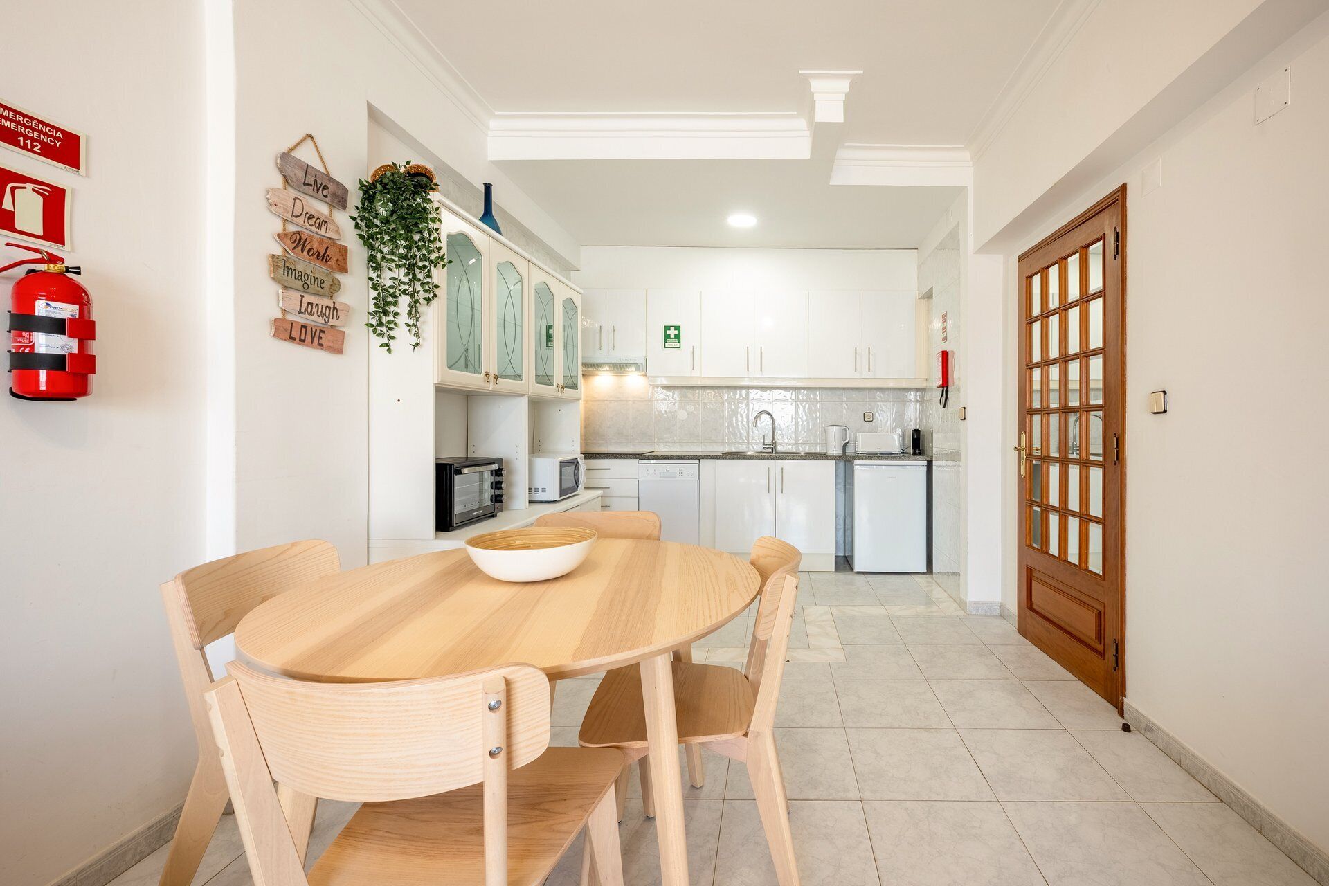 Una cocina con armarios blancos, un microondas y un lavabo. Hay un extintor de incendios en la pared y una puerta con panel de vidrio.