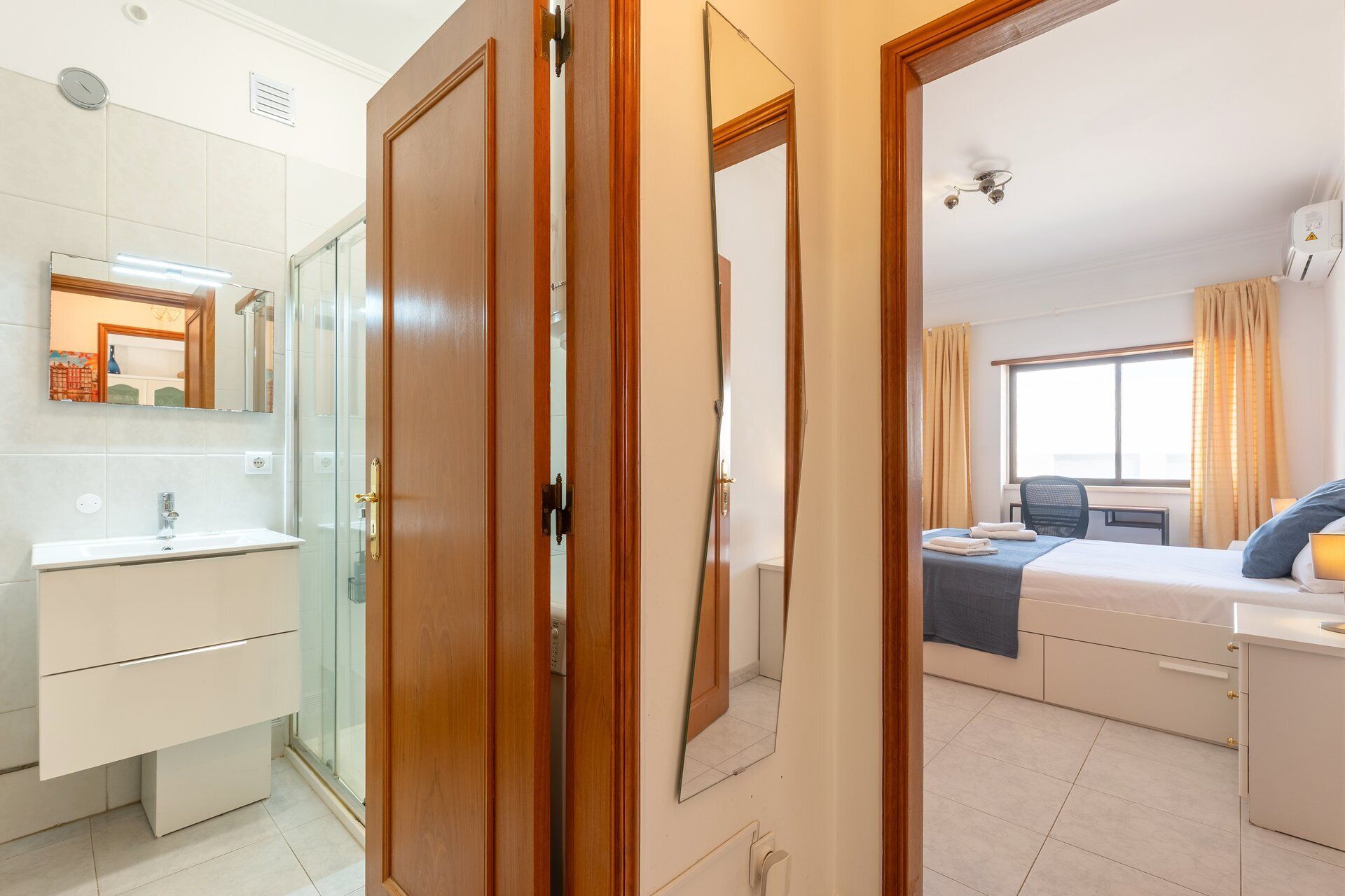 Habitación de hotel con cama, mesita de noche, escritorio, silla, ventana con cortinas, baño con lavamanos, espejo y ducha.