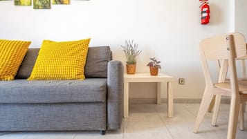 Un salon avec un canapé gris, deux coussins jaunes, une petite table sur laquelle sont posées deux plantes en pot, une chaise en bois et un extincteur fixé au mur.