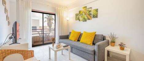 A living room with a grey sofa, yellow cushions, a white coffee table, wicker furniture, a flat-screen TV, and artwork on the wall.