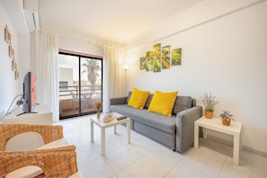 Una sala de estar con un sofá gris, cojines amarillos, una mesa de centro blanca, muebles de mimbre, un televisor de pantalla plana y una obra de arte en la pared.