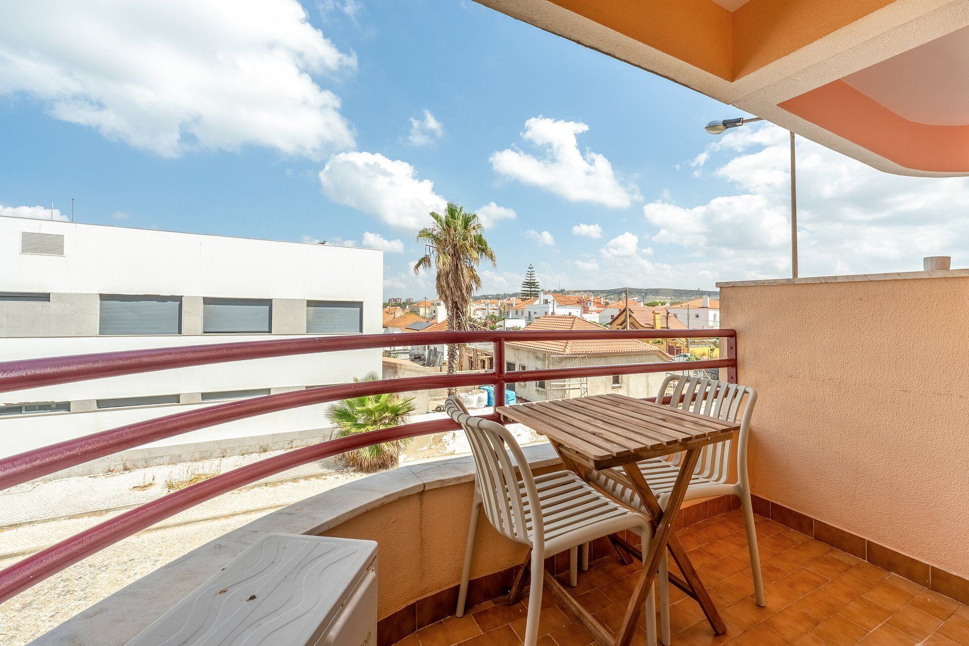 Un balcon avec une table et des chaises en bois, une rambarde en métal, et une vue sur des immeubles et un palmier en arrière-plan.
