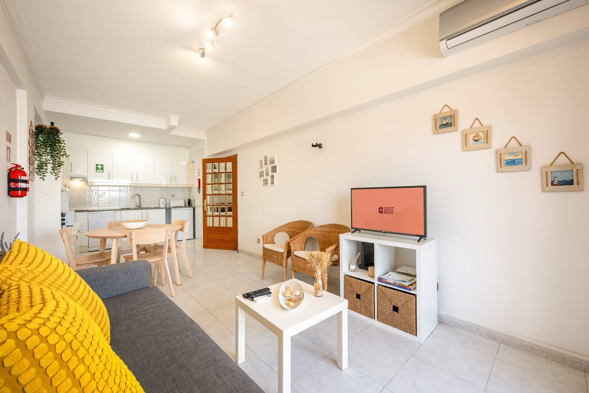 Een moderne woonkamer met een bank, een salontafel en een televisie. Achtergrond is een keuken met witte kasten en een brandblusser aan de muur.