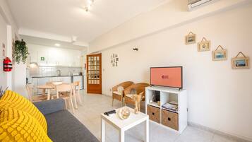Een moderne woonkamer met een bank, een salontafel en een televisie. Achtergrond is een keuken met witte kasten en een brandblusser aan de muur.